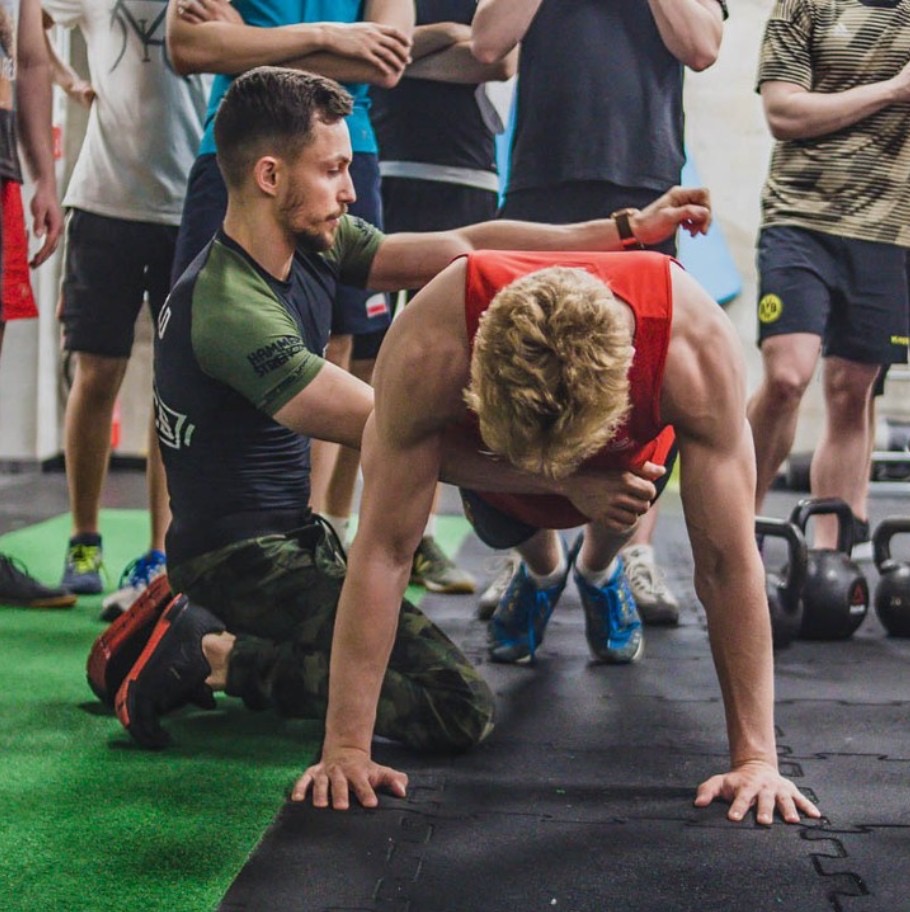 Trener personalny koryguje pozycję klienta podczas ćwiczenia pompki w siłowni. W tle grupa obserwujących, kettlebells i zielona podłoga.