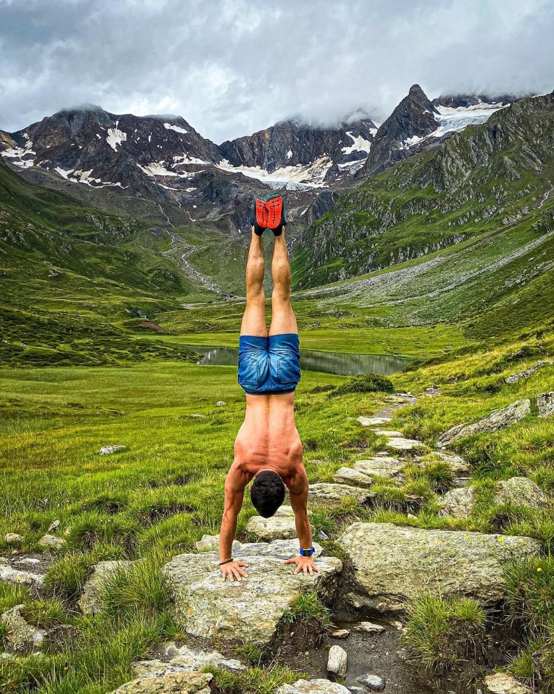 Mężczyzna w pozycji stania na rękach na tle górskiego krajobrazu z jeziorem. Ćwiczenie na kamieniu, boso, w krótkich spodenkach i sportowych butach.