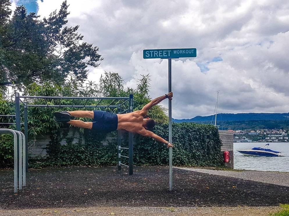 Mężczyzna w pozycji 'flaga' na słupie z tabliczką 'Street Workout' na tle jeziora i zieleni. Widoczne drabinki do ćwiczeń i łódź na wodzie.