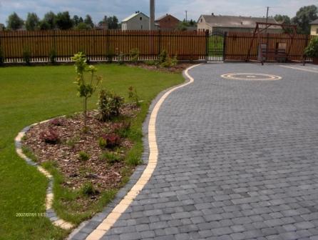 Aranżacja ogrodu z wybrukowanym podjazdem z kostki w odcieniach szarości, obrzeżem z jasnego kamienia i rabatą wysypaną korą, na której rosną młode drzewka i krzewy