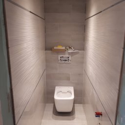 F.H.U. PYRAMID JacekPyra - Nowoczesna toaleta podwieszana w wąskiej wnęce wyłożonej beżowymi płytkami z delikatną falistą strukturą, z widocznymi elementami instalacji wodnej i elektrycznej oraz półką z akcesoriami.