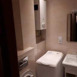 F.H.U. PYRAMID JacekPyra - Kompaktowa łazienka z pralką ładowaną od góry, wiszącym piecykiem gazowym z odprowadzeniem spalin i beżowymi płytkami ściennymi.