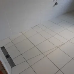 Białe, kwadratowe płytki podłogowe i ścienne w kabinie prysznicowej z odpływem liniowym; widoczny fragment surowej ściany z cegły.