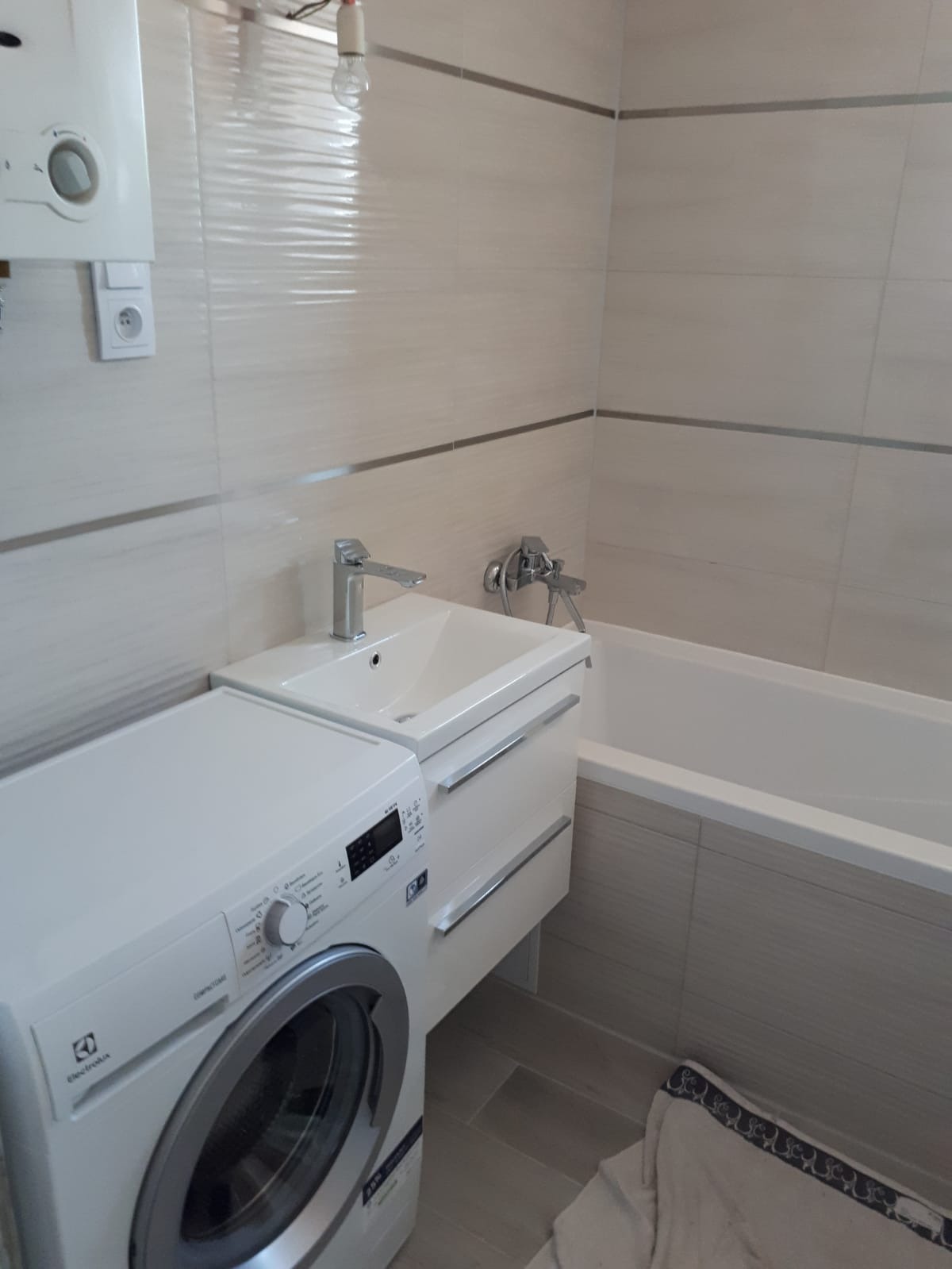 Jasna łazienka z umywalką na szafce z szufladami, pralką Electrolux CompactGo obok, białą wanną i beżowymi płytkami z poziomymi paskami; widoczny także bojler na ścianie.