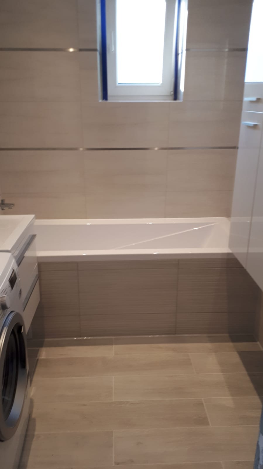 Jasna łazienka z beżowymi płytkami, białą wanną zabudowaną płytkami, pralką i oknem z niebieską ramą.