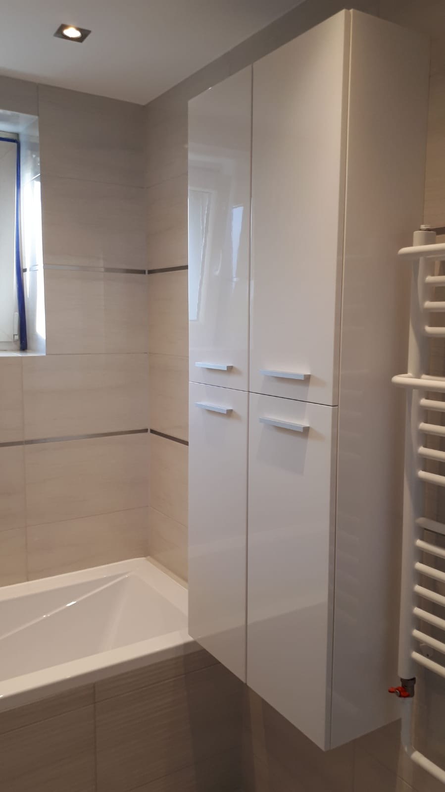 Biała, lakierowana szafka łazienkowa z czterema drzwiczkami i podłużnymi uchwytami, zamontowana nad wanną w łazience wykończonej beżowymi płytkami z metalowymi listwami dekoracyjnymi.