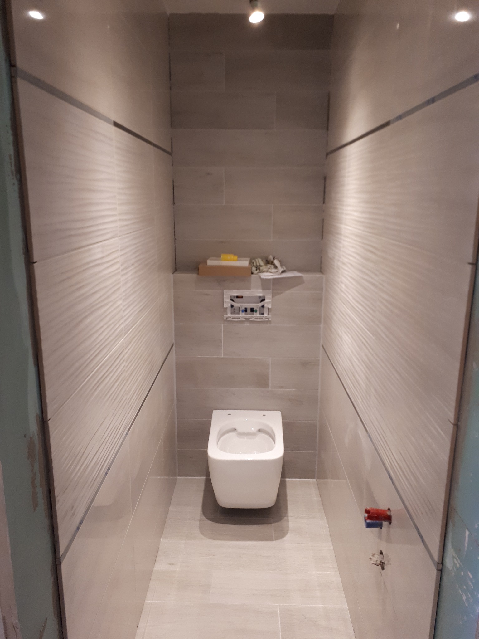 Nowoczesna toaleta podwieszana w wąskiej wnęce wyłożonej beżowymi płytkami z delikatną falistą strukturą, z widocznymi elementami instalacji wodnej i elektrycznej oraz półką z akcesoriami.