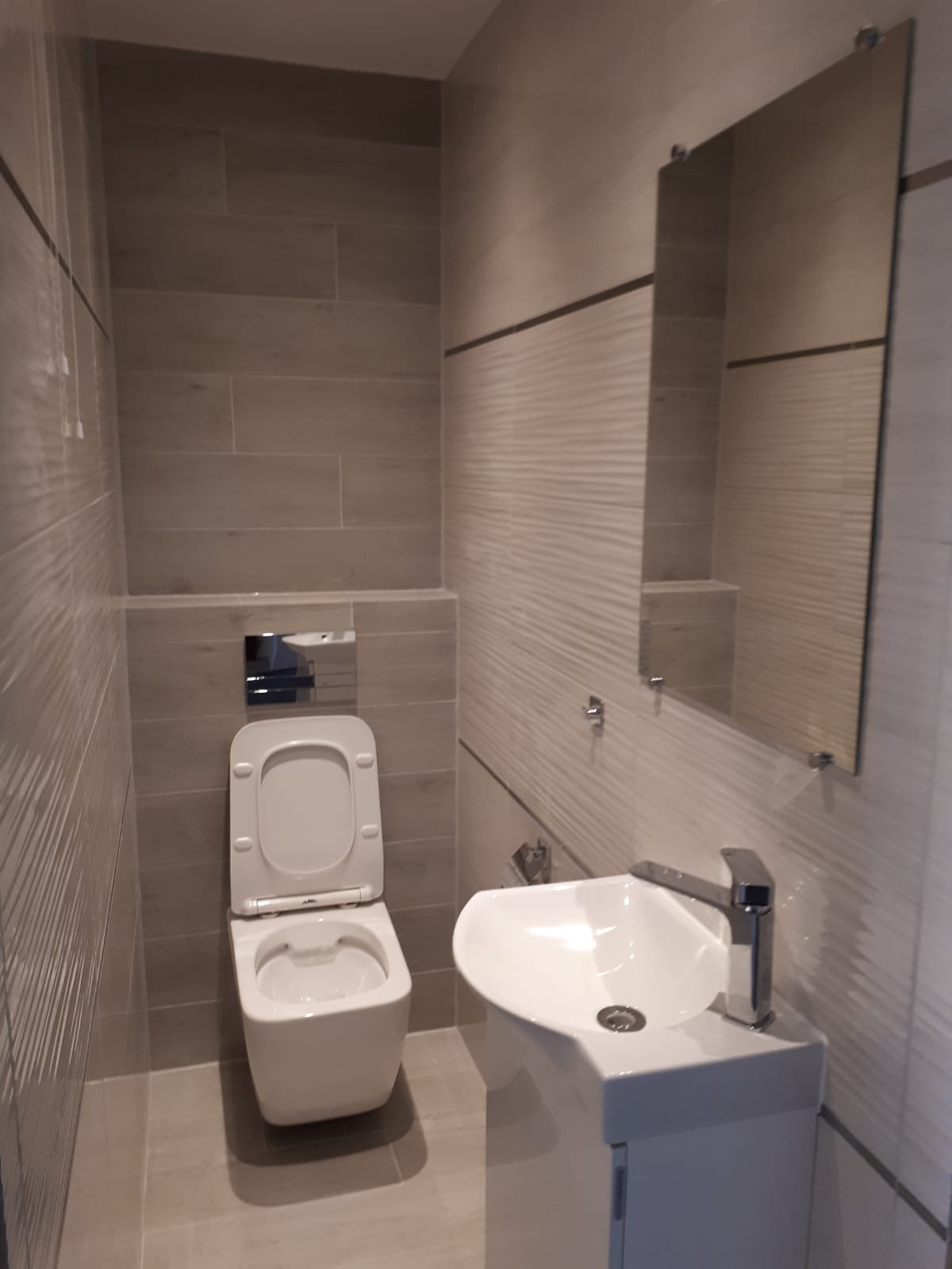 Nowoczesna łazienka po remoncie, z wiszącą toaletą z podtynkową spłuczką, umywalką na szafce i dużym lustrem, wykończona płytkami w odcieniach szarości z delikatnym wzorem.