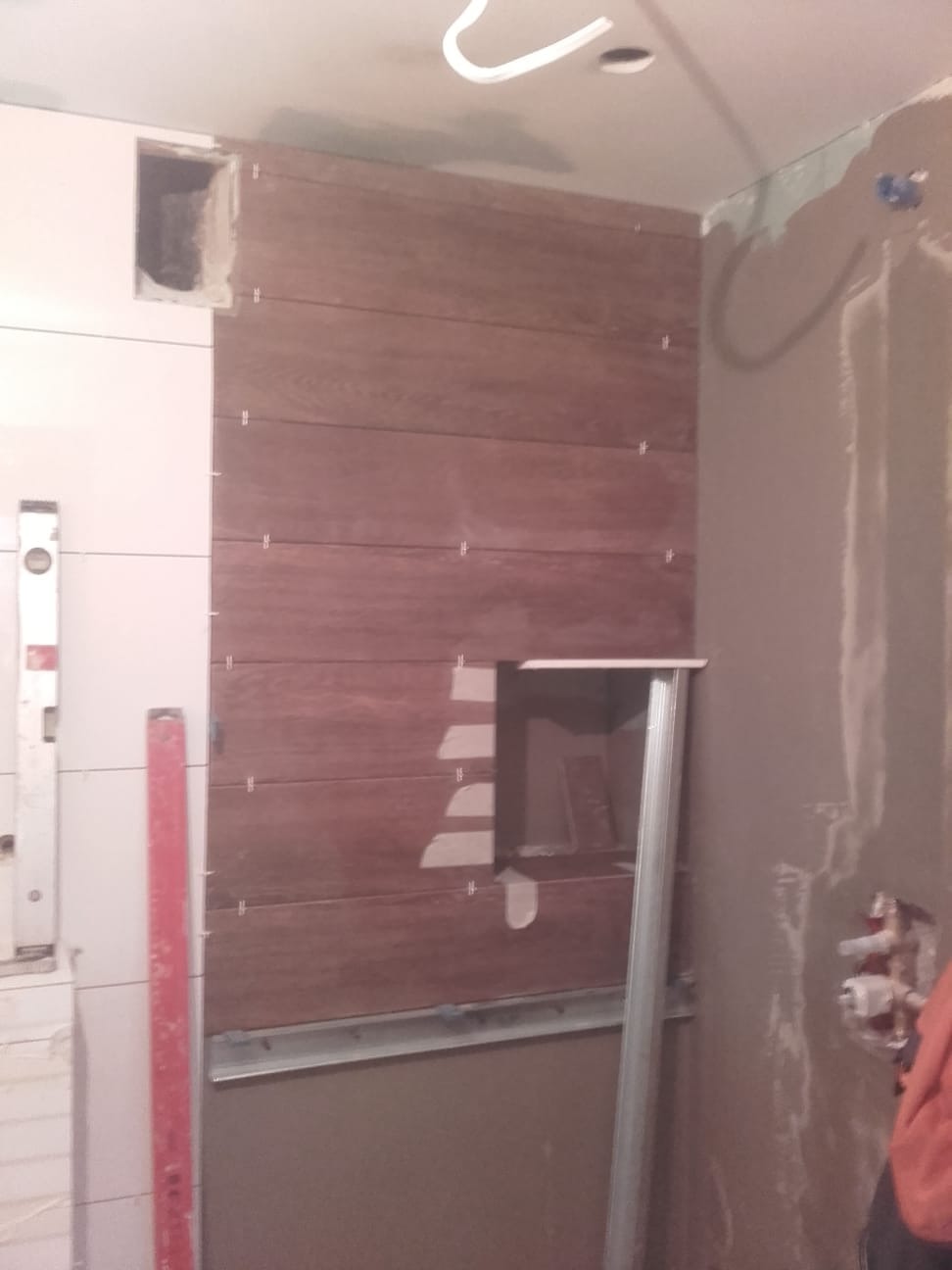 Montaż drewnopodobnych płytek na ścianie w łazience, widoczne poziomice i metalowe profile pomocnicze, ściana w trakcie wykończenia, częściowo otynkowana.