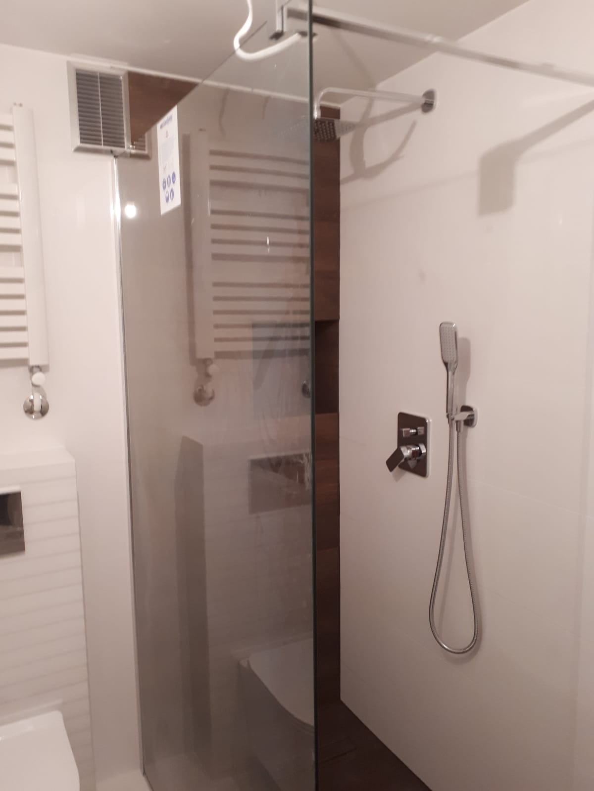 Nowoczesna kabina prysznicowa z szklaną ścianką, deszczownicą i baterią podtynkową w łazience z białymi płytkami i grzejnikiem drabinkowym.