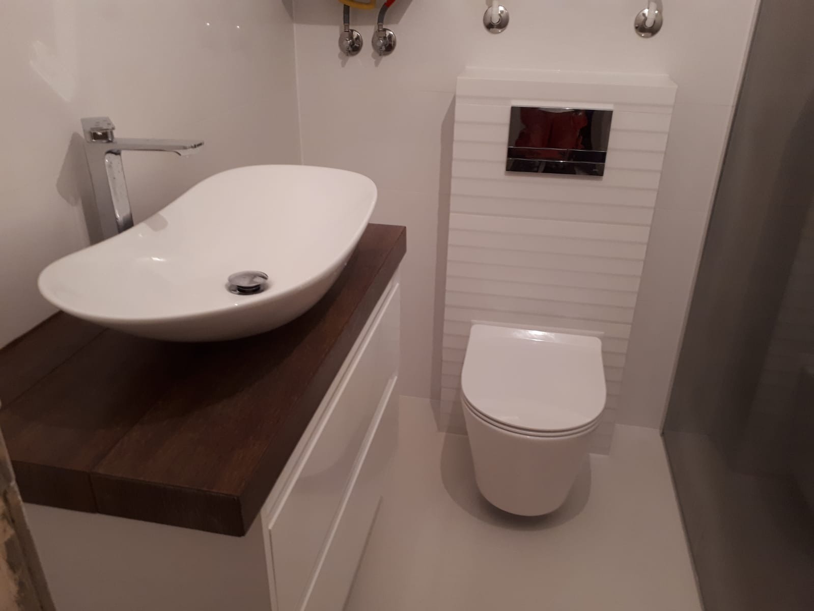 Nowoczesna łazienka z owalną umywalką nablatową na ciemnym drewnianym blacie, wisząca toaleta z ukrytym systemem spłukiwania i białe płytki na ścianach.