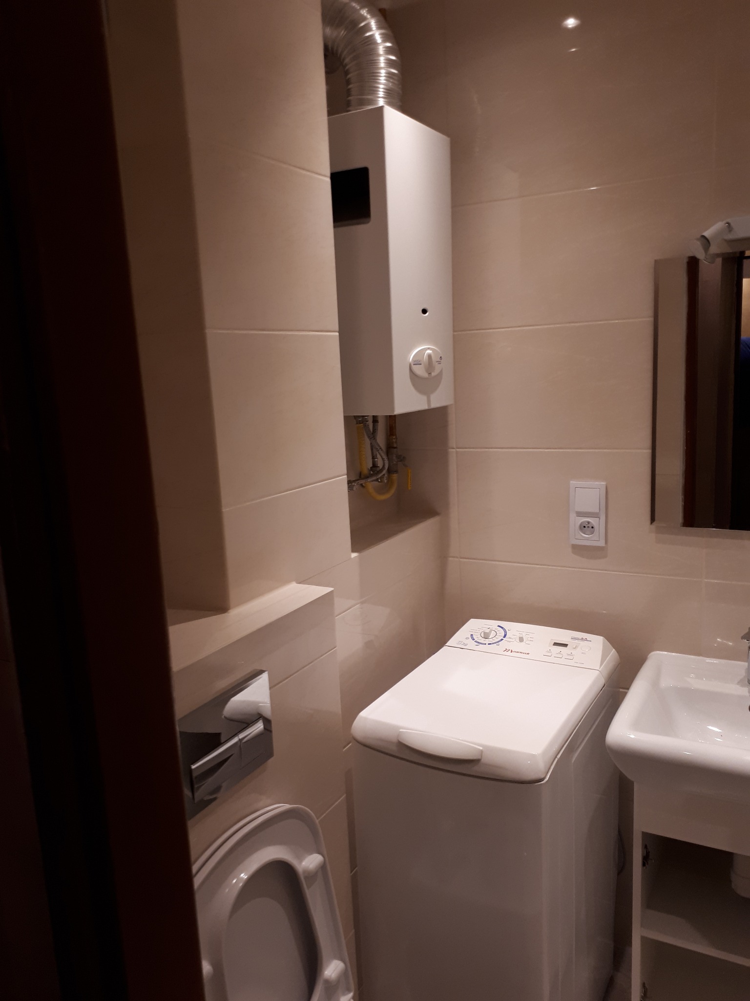 Kompaktowa łazienka z pralką ładowaną od góry, wiszącym piecykiem gazowym z odprowadzeniem spalin i beżowymi płytkami ściennymi.