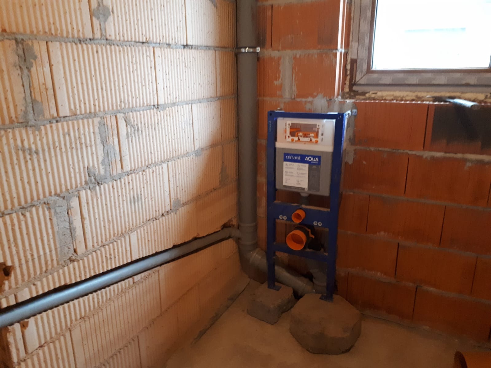 Stelaż podtynkowy WC Cersanit Aqua zamontowany w surowym pomieszczeniu z ceglanymi ścianami i widocznymi rurami kanalizacyjnymi przed zabudową.