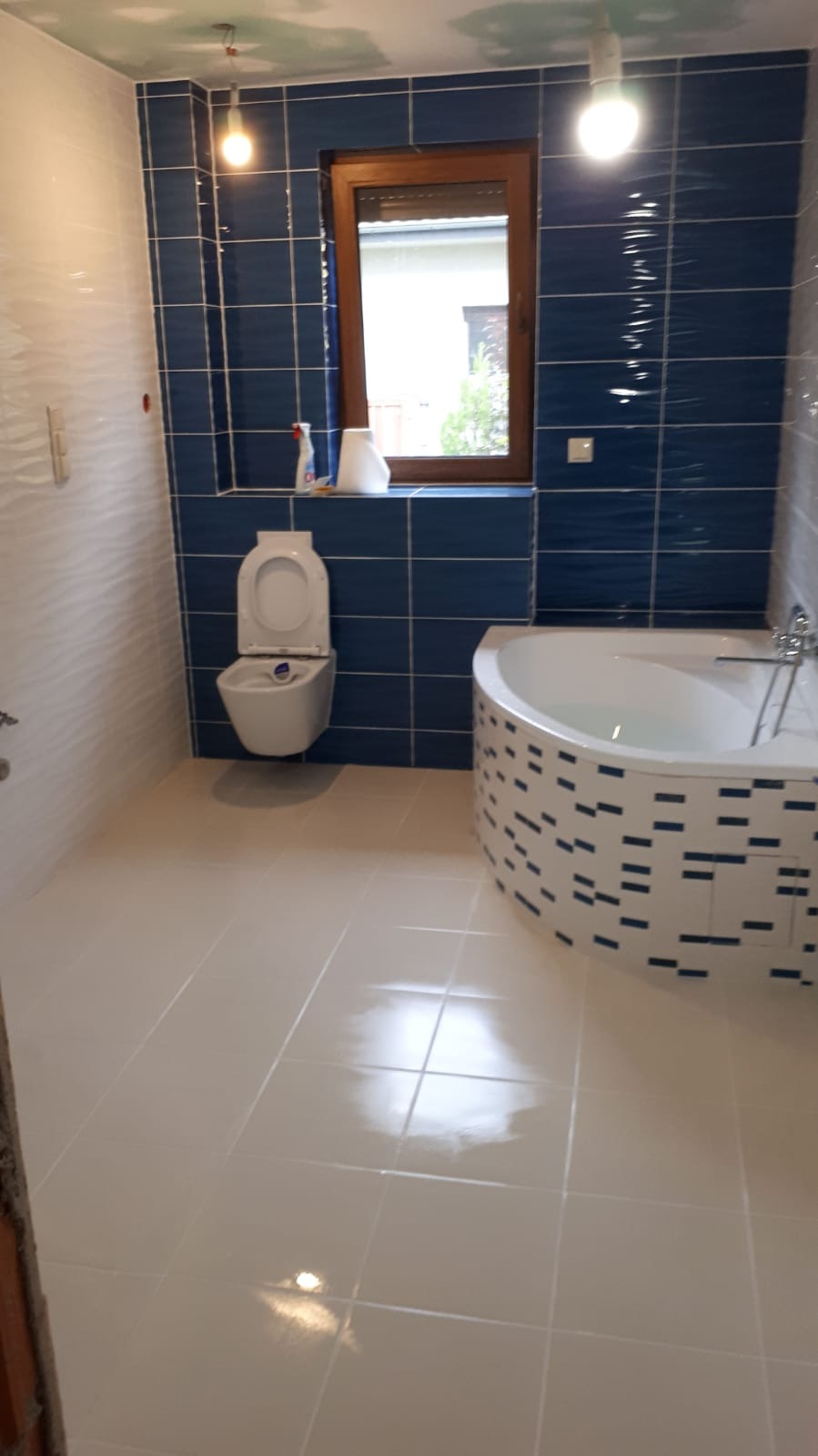 Wykończona łazienka z niebieskimi płytkami na ścianach, białą wanną z mozaiką, toaletą pod oknem i jasnymi płytkami podłogowymi. Sufit w trakcie wykończenia, widoczne przewody elektryczne.