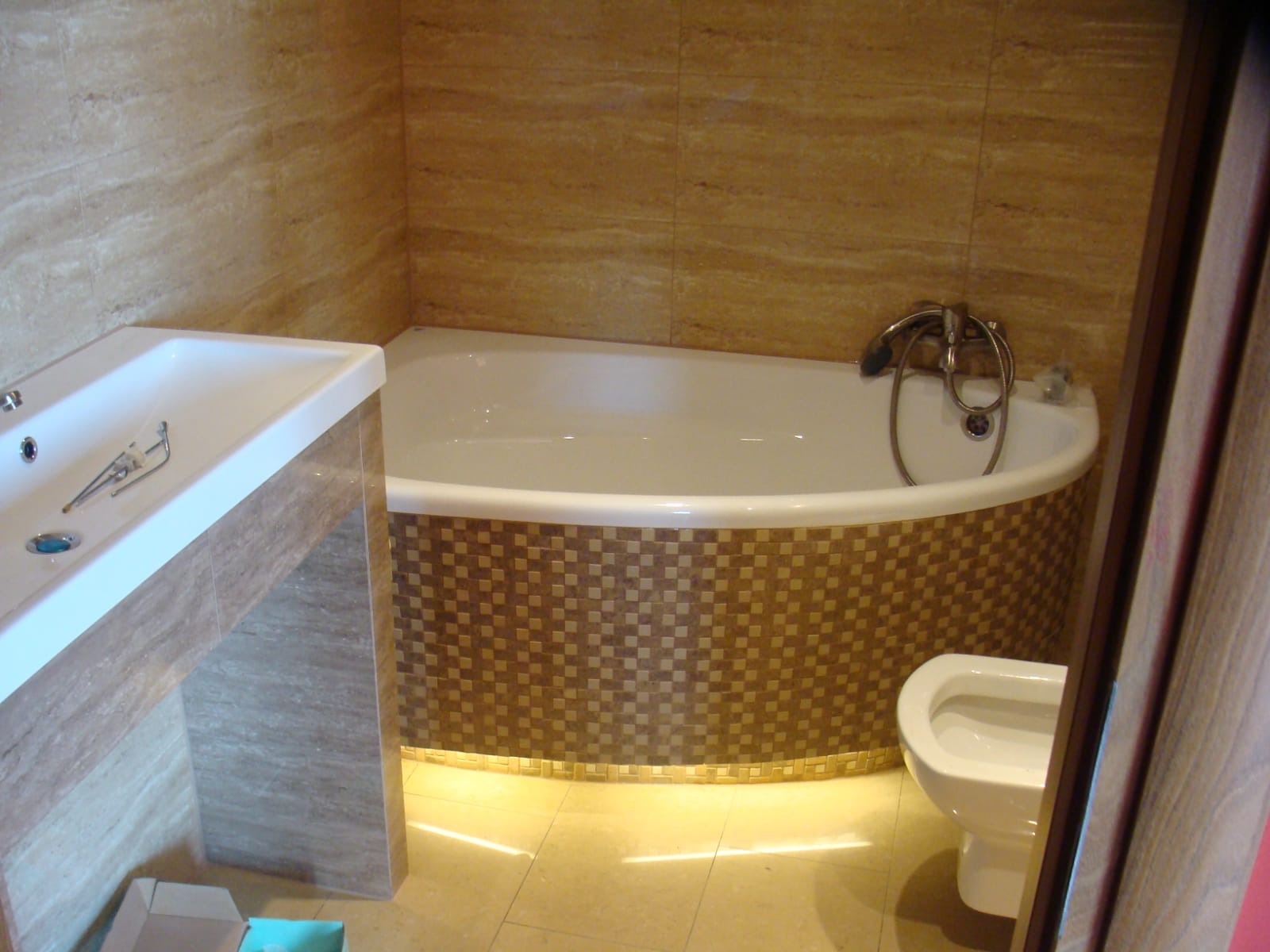 Wykończona łazienka z beżowymi płytkami imitującymi trawertyn na ścianach, owalna wanna z mozaiką na obudowie podświetloną od dołu, umywalka nablatowa na kamiennym blacie i biała toaleta.