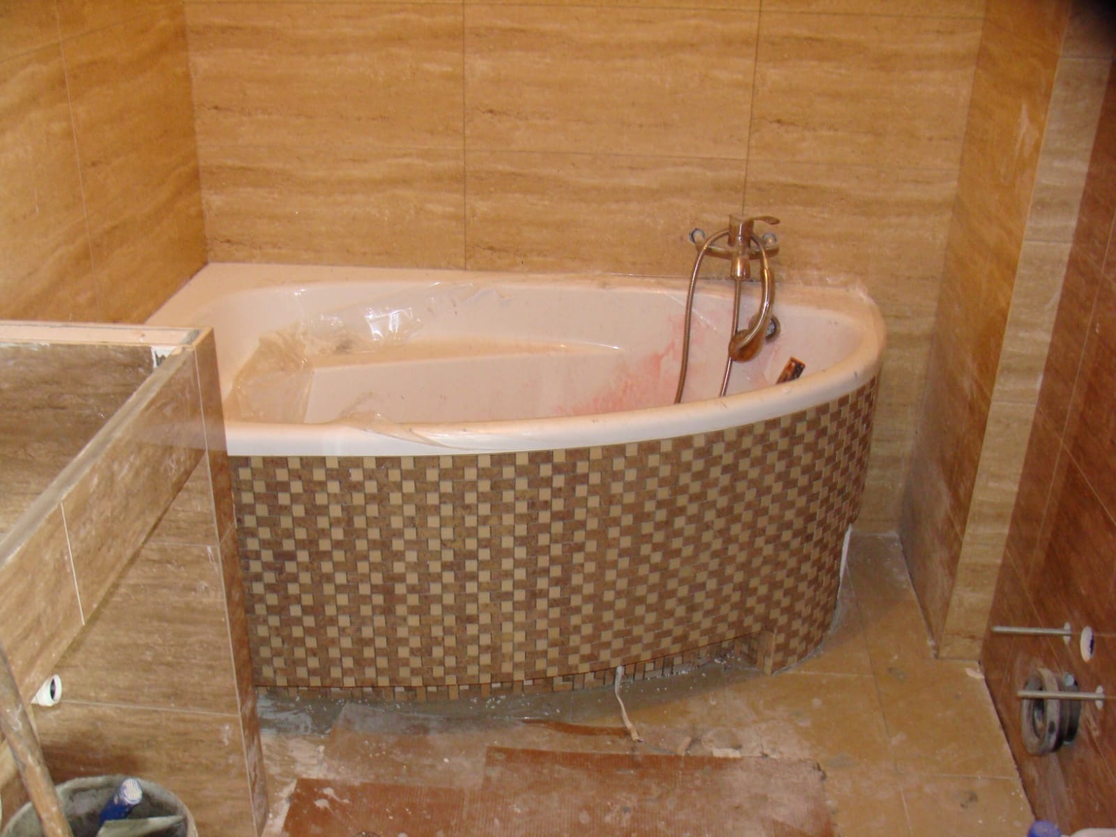 Boczny widok na wannę z mozaikową obudową w trakcie remontu łazienki, zabezpieczona folią, z widocznymi narzędziami i fragmentami płytek na podłodze.