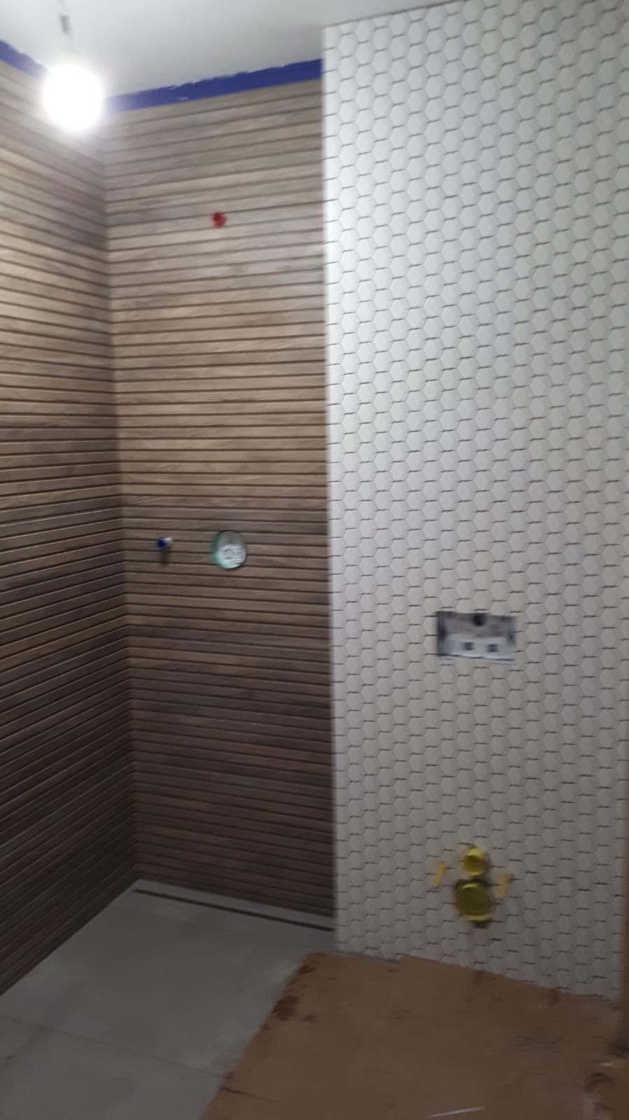 Wnętrze łazienki w trakcie remontu: ściana wyłożona drewnopodobnymi panelami poziomymi oraz ściana z białej mozaiki heksagonalnej, widoczne przygotowane otwory na instalacje i zabezpieczenia folią.