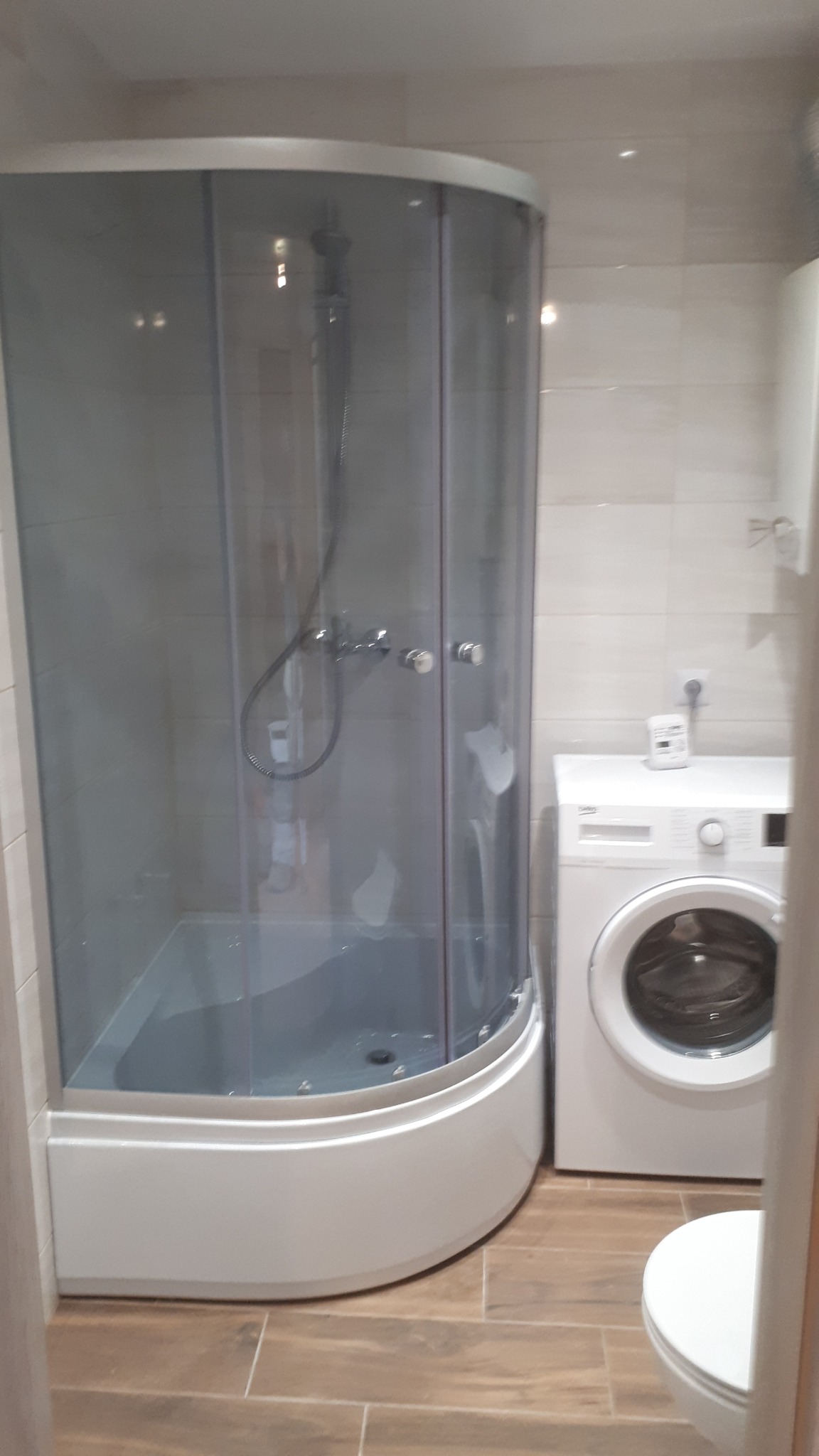 Kabinowa narożna kabina prysznicowa z chromowaną armaturą w łazience wykończonej płytkami imitującymi drewno, obok pralka.