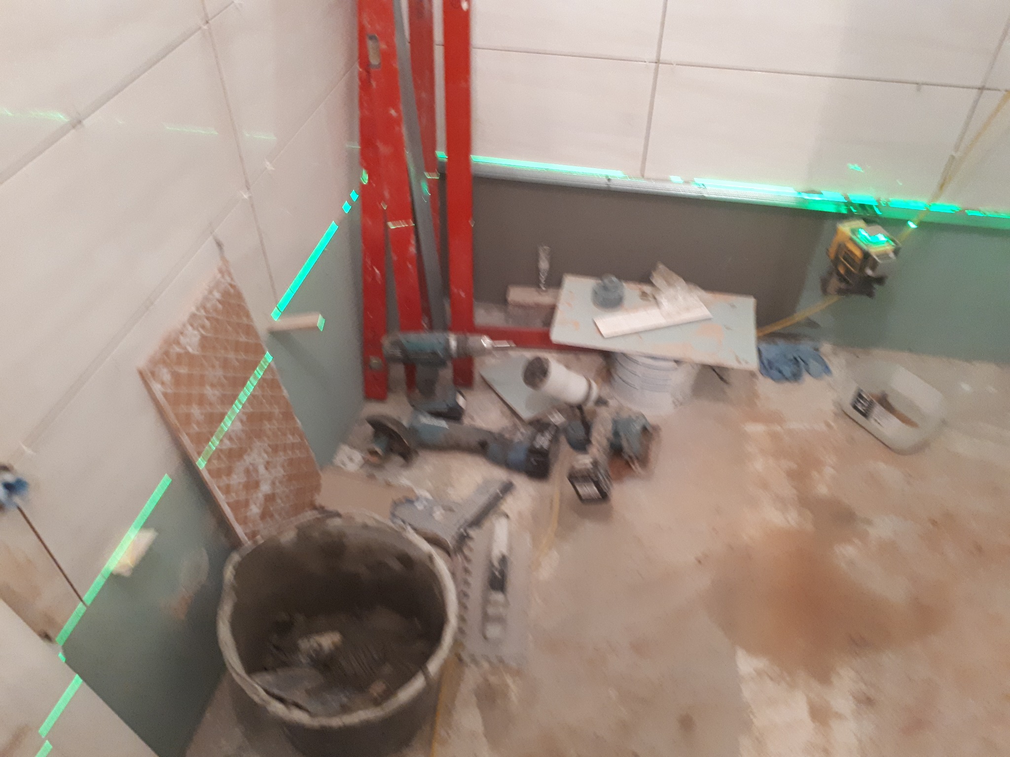 Niedokończona łazienka z ułożonymi białymi płytkami na ścianach, narzędzia i wiadro z zaprawą na podłodze, poziomica laserowa wyznacza linie.
