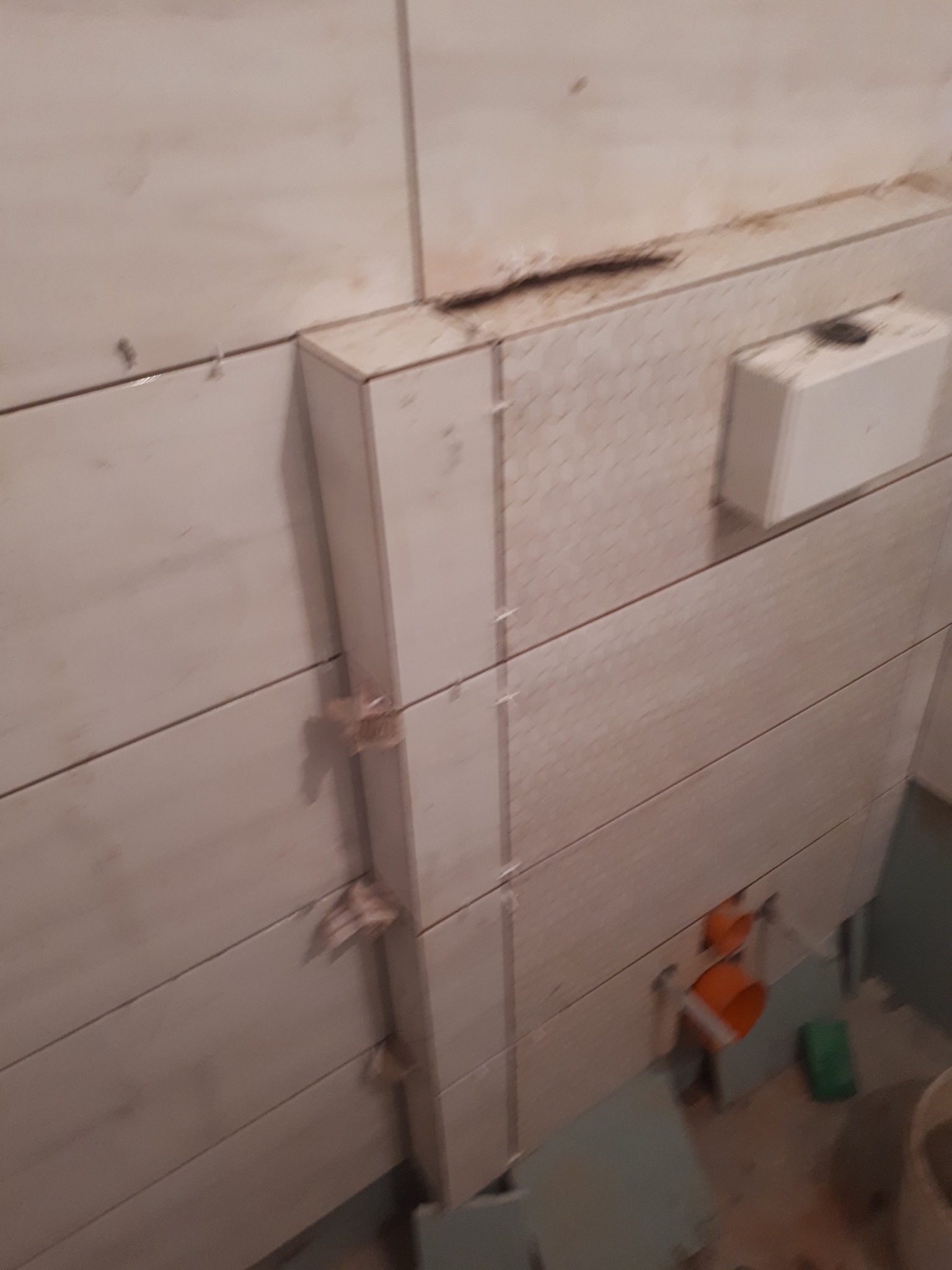 Ściana w trakcie układania płytek, widoczne plastikowe krzyżyki dystansowe, fragment instalacji sanitarnej i niedokończone fugi.