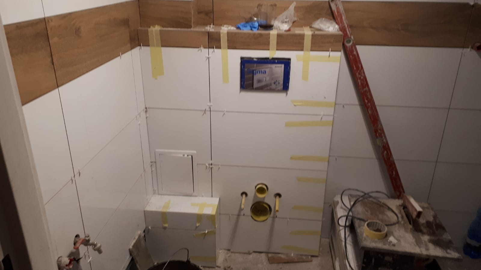 Ściana łazienki w trakcie układania białych płytek z drewnopodobnym pasem u góry, widoczne żółte taśmy, poziomica oparta o ścianę i narzędzia glazurnicze na podłodze.