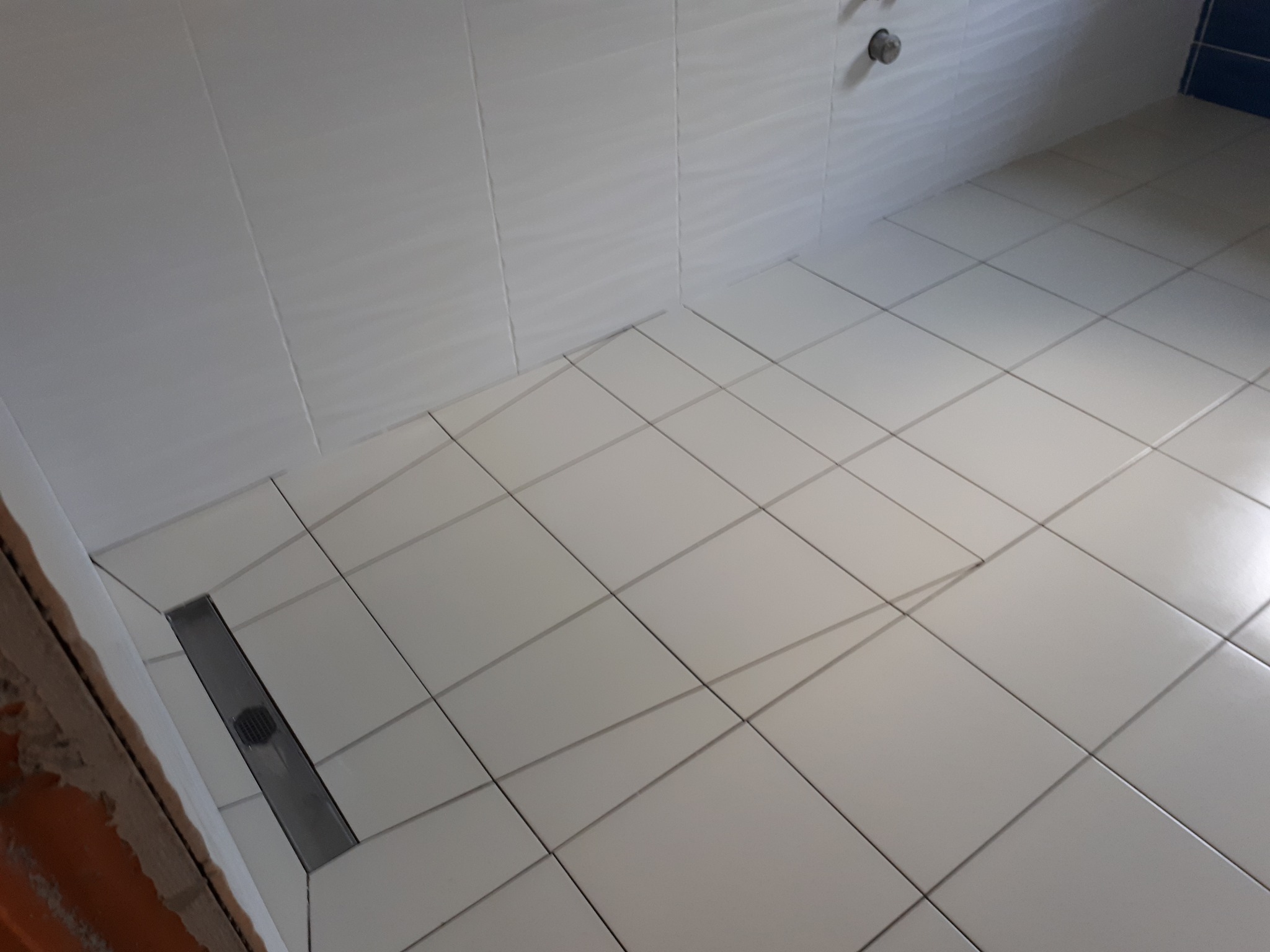 Białe, kwadratowe płytki podłogowe i ścienne w kabinie prysznicowej z odpływem liniowym; widoczny fragment surowej ściany z cegły.