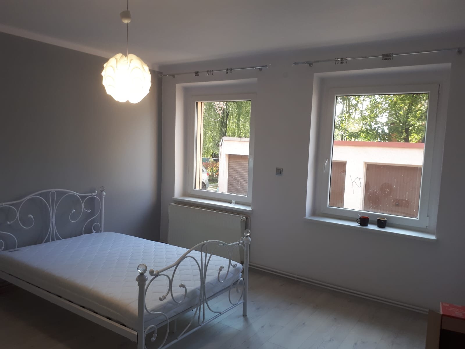 Pomalowane na szaro wnętrze pokoju z białym, metalowym łóżkiem, widokiem z okna na garaże i wiszącą lampą w kształcie kwiatu.