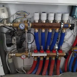 Rozdzielacz ogrzewania podłogowego w szafce, widoczne rury doprowadzające wodę w kolorze niebieskim i odprowadzające w kolorze czerwonym, manometry, termostaty Salus i pompa obiegowa.