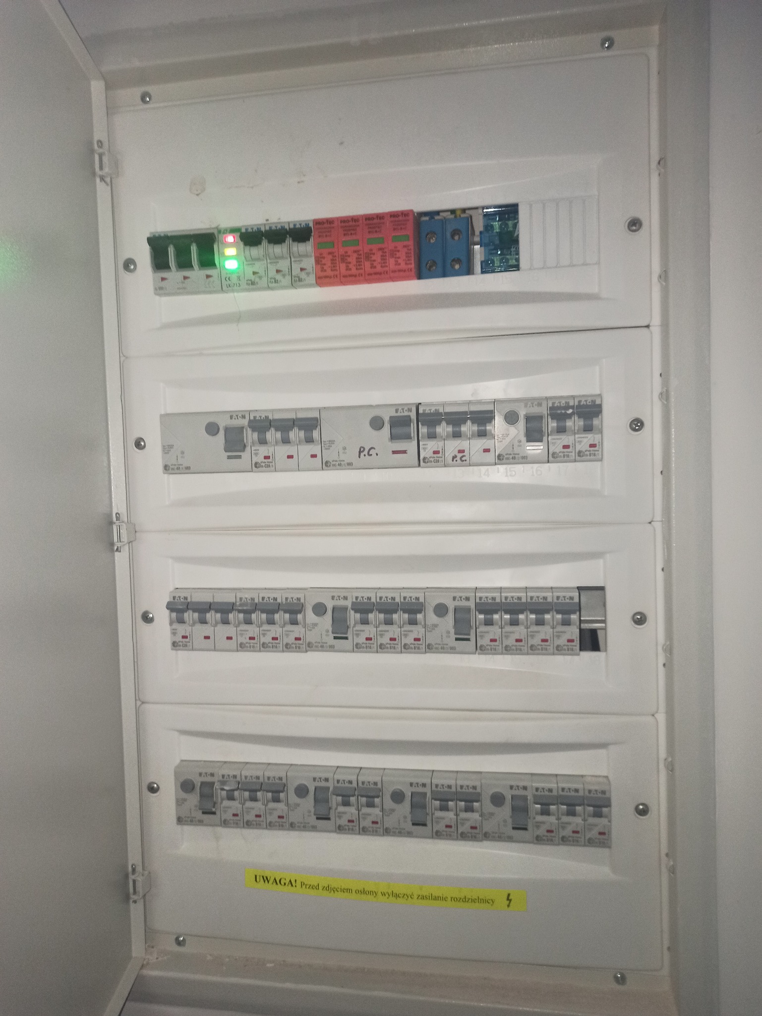 Rozdzielnica elektryczna w białej obudowie, widoczne zabezpieczenia różnicowoprądowe, bezpieczniki automatyczne Eaton z oznaczeniami P.C. i żółta naklejka ostrzegawcza.