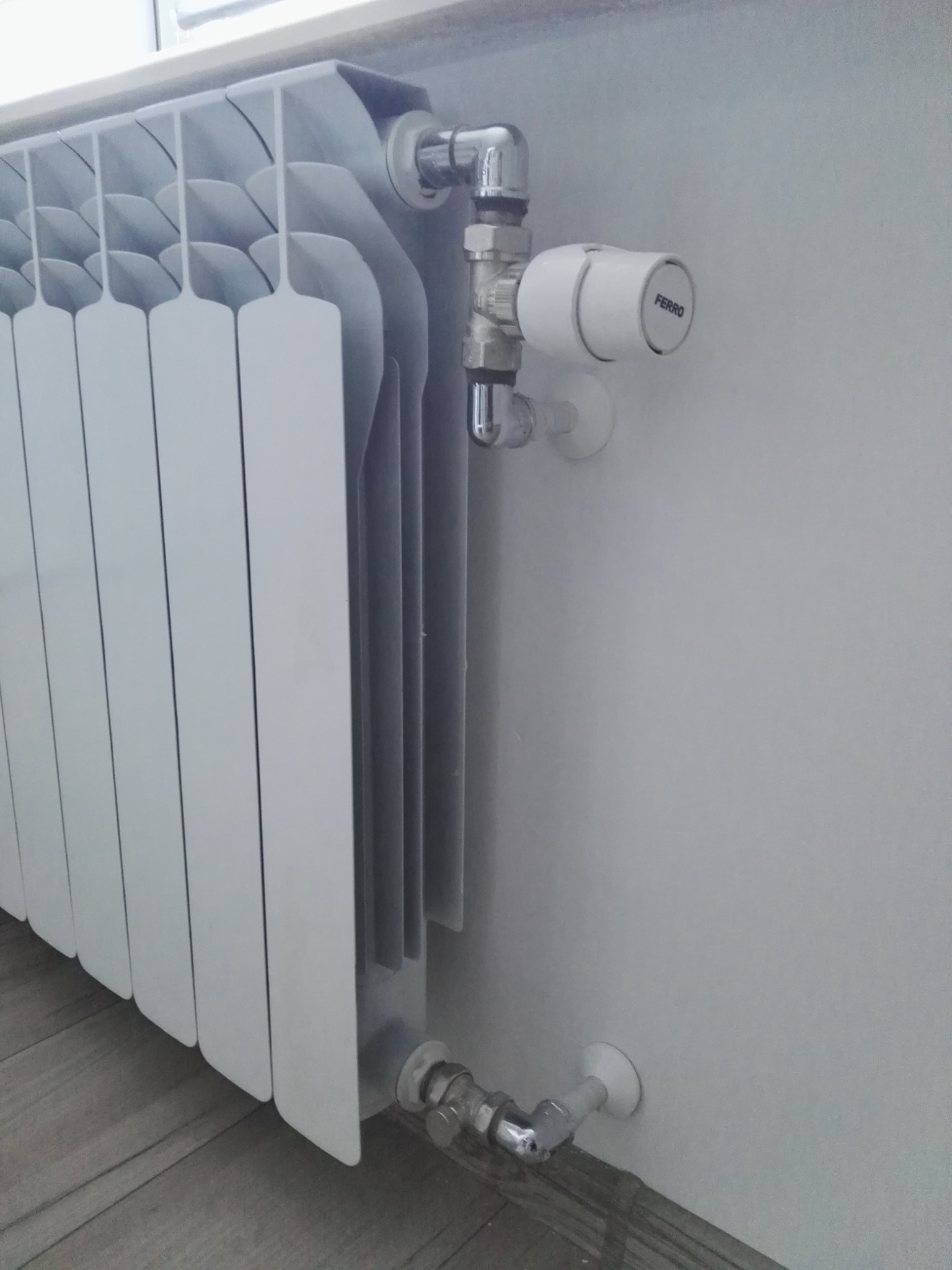 Biały, aluminiowy grzejnik panelowy z termostatem Ferro zamontowany przy ścianie, widoczne przyłącza wodne.