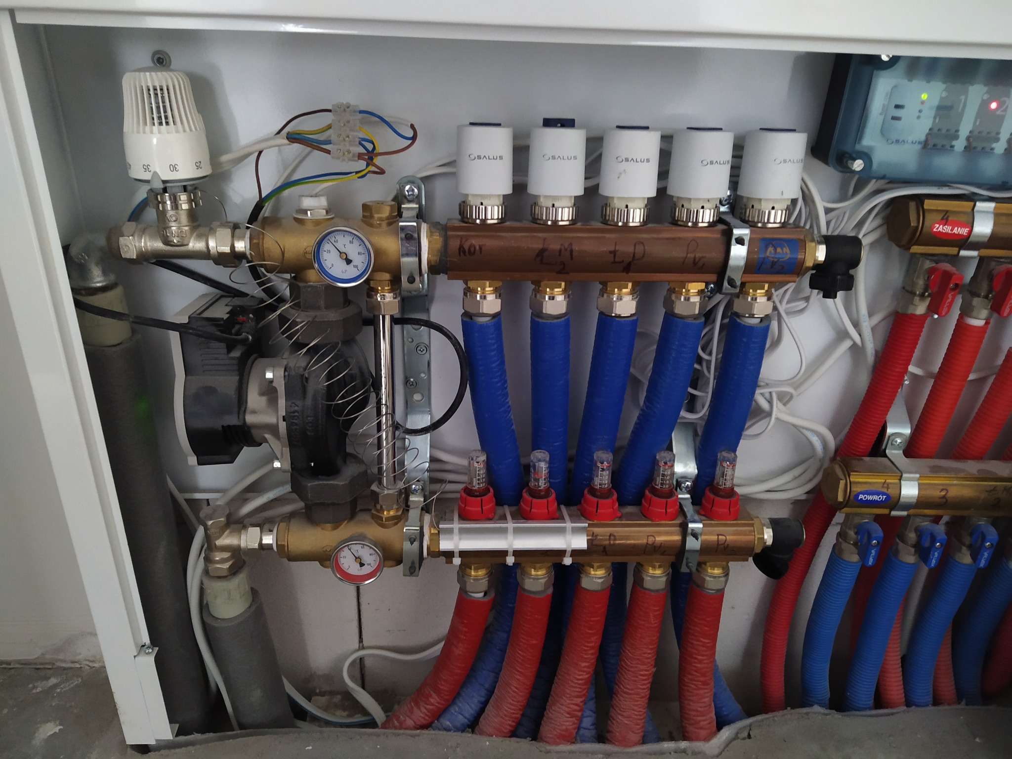 Rozdzielacz ogrzewania podłogowego w szafce, widoczne rury doprowadzające wodę w kolorze niebieskim i odprowadzające w kolorze czerwonym, manometry, termostaty Salus i pompa obiegowa.