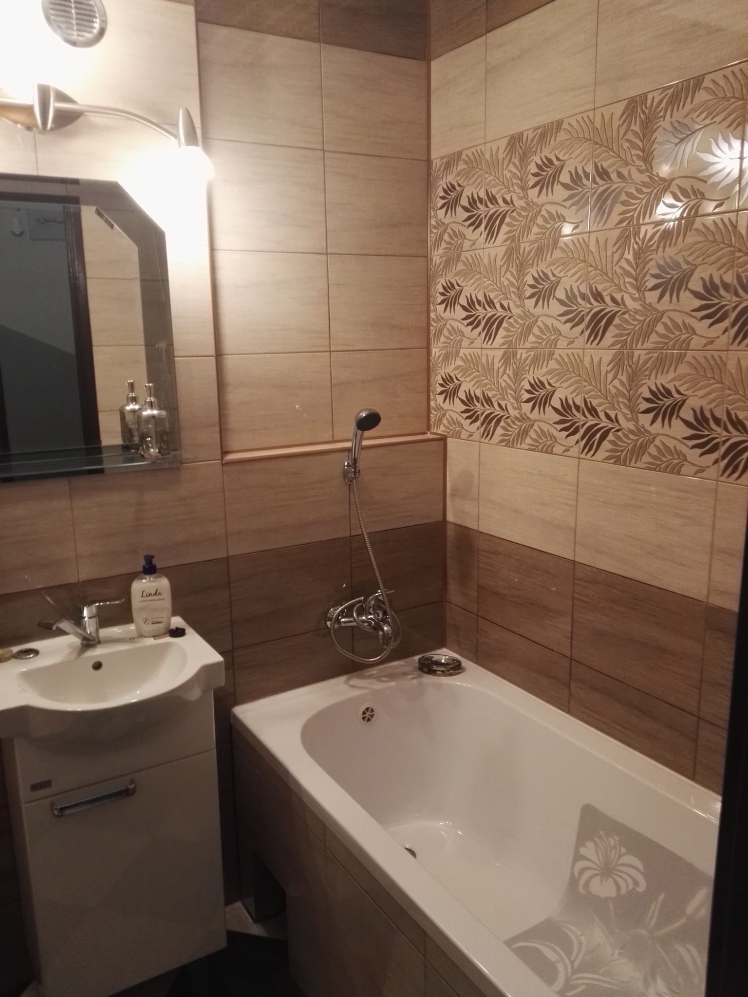 Umywalka z szafką, wanna z antypoślizgową naklejką w kwiaty i chromowana bateria prysznicowa na tle beżowych płytek z motywem liści.