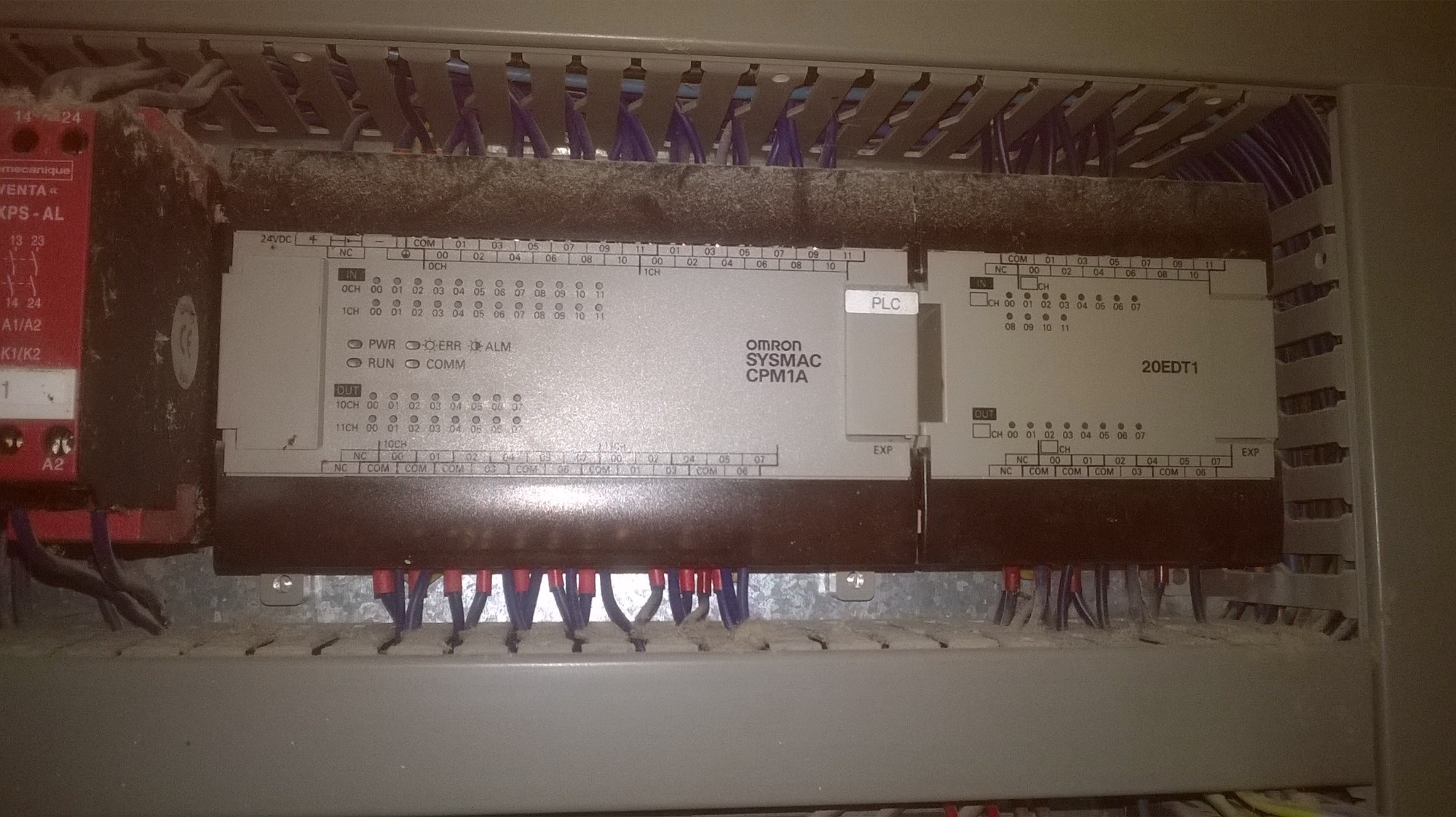 Sterownik PLC Omron Sysmac CPM1A w szafie sterowniczej, widoczne liczne przewody i moduły rozszerzeń, całość lekko zakurzona.