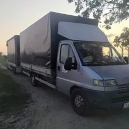 Srebrny bus z przyczepą plandekową, zaparkowany na polnej drodze o zmierzchu, widok z boku.