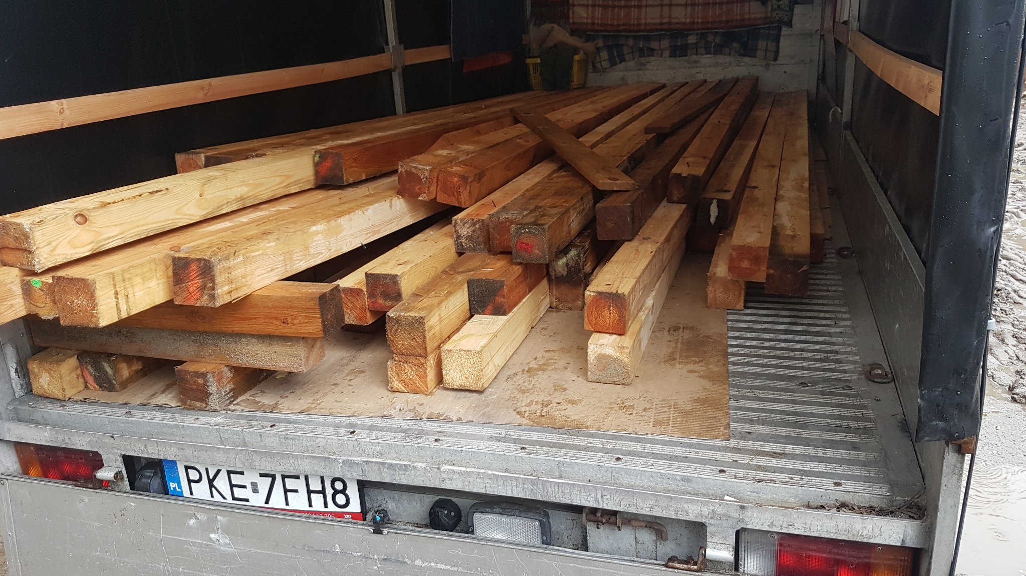 Kwadratowe drewniane belki ułożone w przestrzeni ładunkowej samochodu dostawczego z widoczną tablicą rejestracyjną.