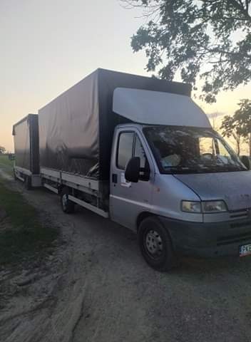 Srebrny bus z przyczepą plandekową, zaparkowany na polnej drodze o zmierzchu, widok z boku.