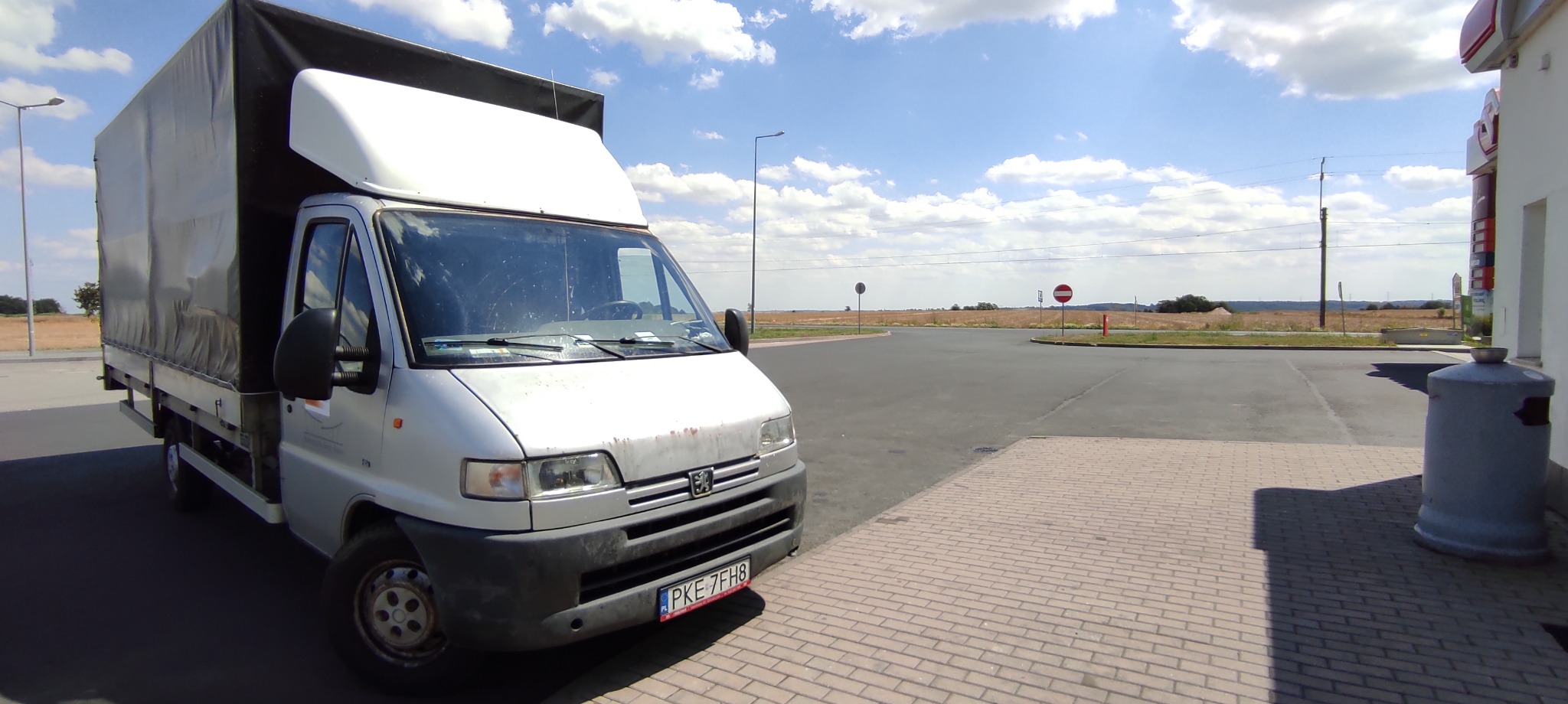 Biały samochód dostawczy z czarną plandeką, zaparkowany na kostce brukowej przy skrzyżowaniu dróg w słoneczny dzień.