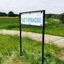 Studio graficzne MBSO - Szyld Lokalnej Organizacji Turystycznej Gminy Sztutowo z nazwą 'Kąty Rybackie' na czarnym stelażu, umieszczony na tle łąki i ścieżki. Odświeżony wygląd.