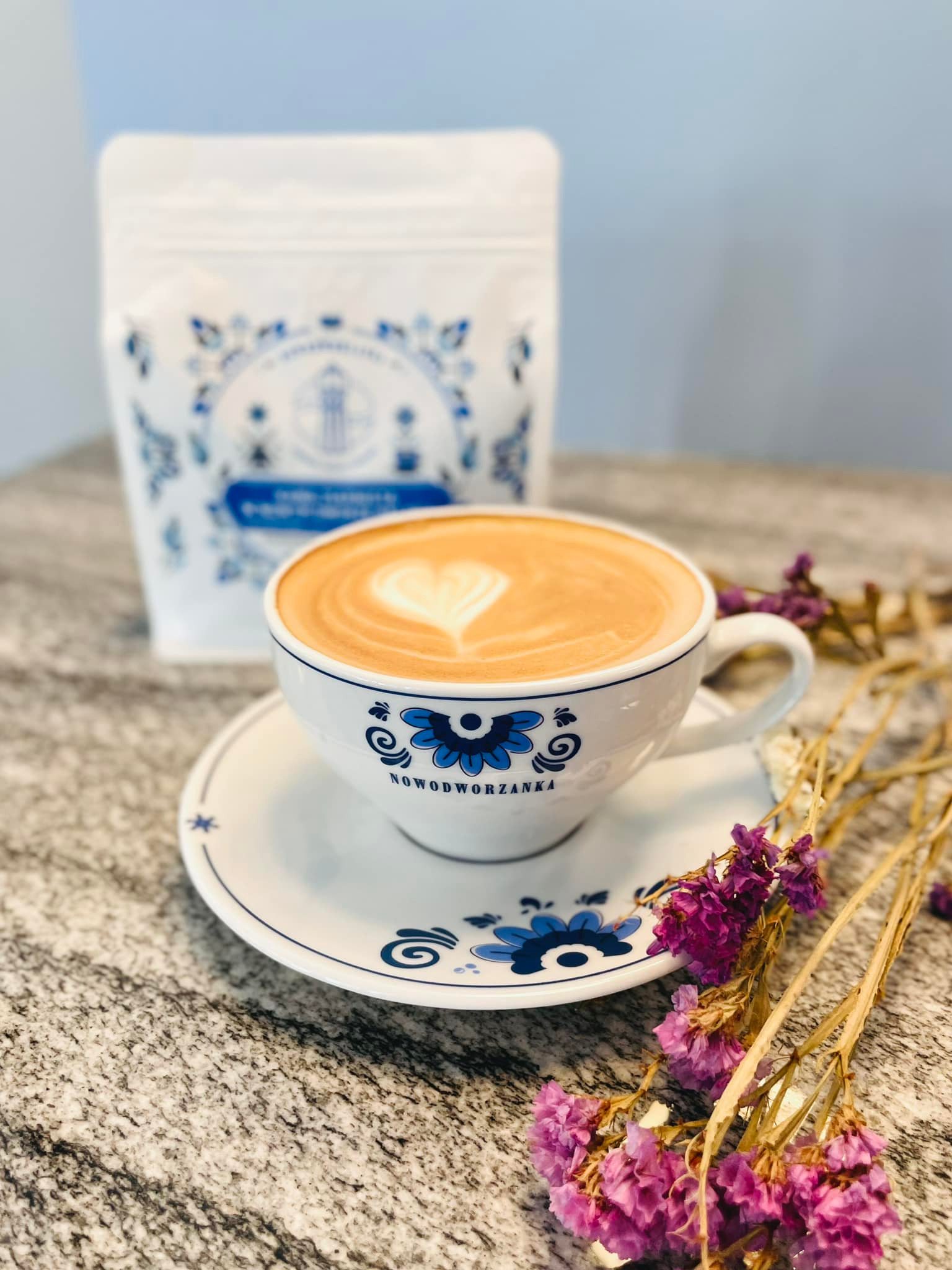 Filiżanka kawy latte z wzorem serca na piance, obok opakowanie kawy z niebieskim nadrukiem kwiatowym i suszone kwiaty, na marmurowym blacie.
