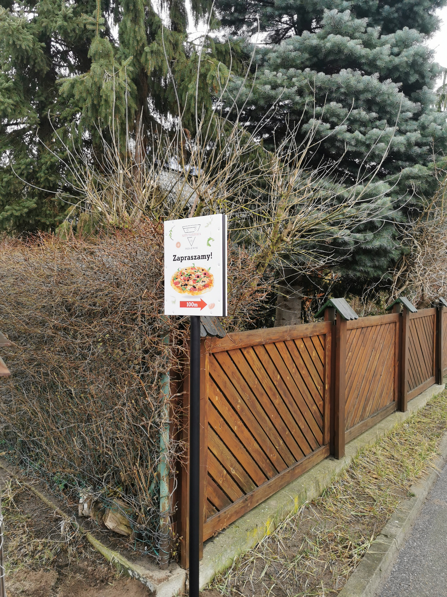 Tablica reklamowa z napisem 'Zapraszamy!' i grafiką pizzy, umieszczona na słupku przed drewnianym płotem i krzewami, wskazująca odległość 100m do pizzerii Sztutozzeria.