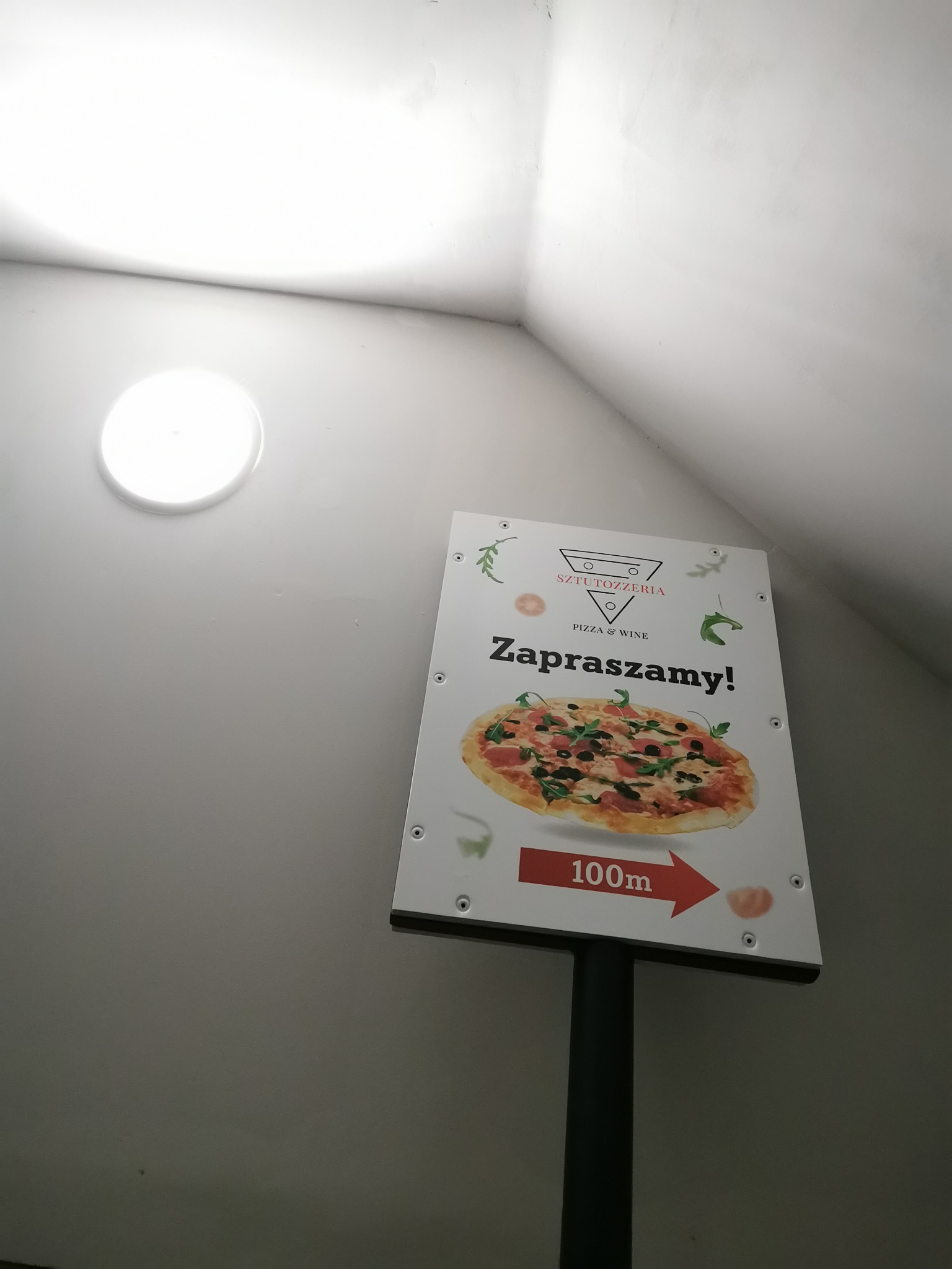 Szyld reklamowy pizzerii Sztutozzeria z grafiką pizzy i napisem 'Zapraszamy!', umieszczony na słupku, wskazujący odległość 100m, sfotografowany w studiu przed montażem.
