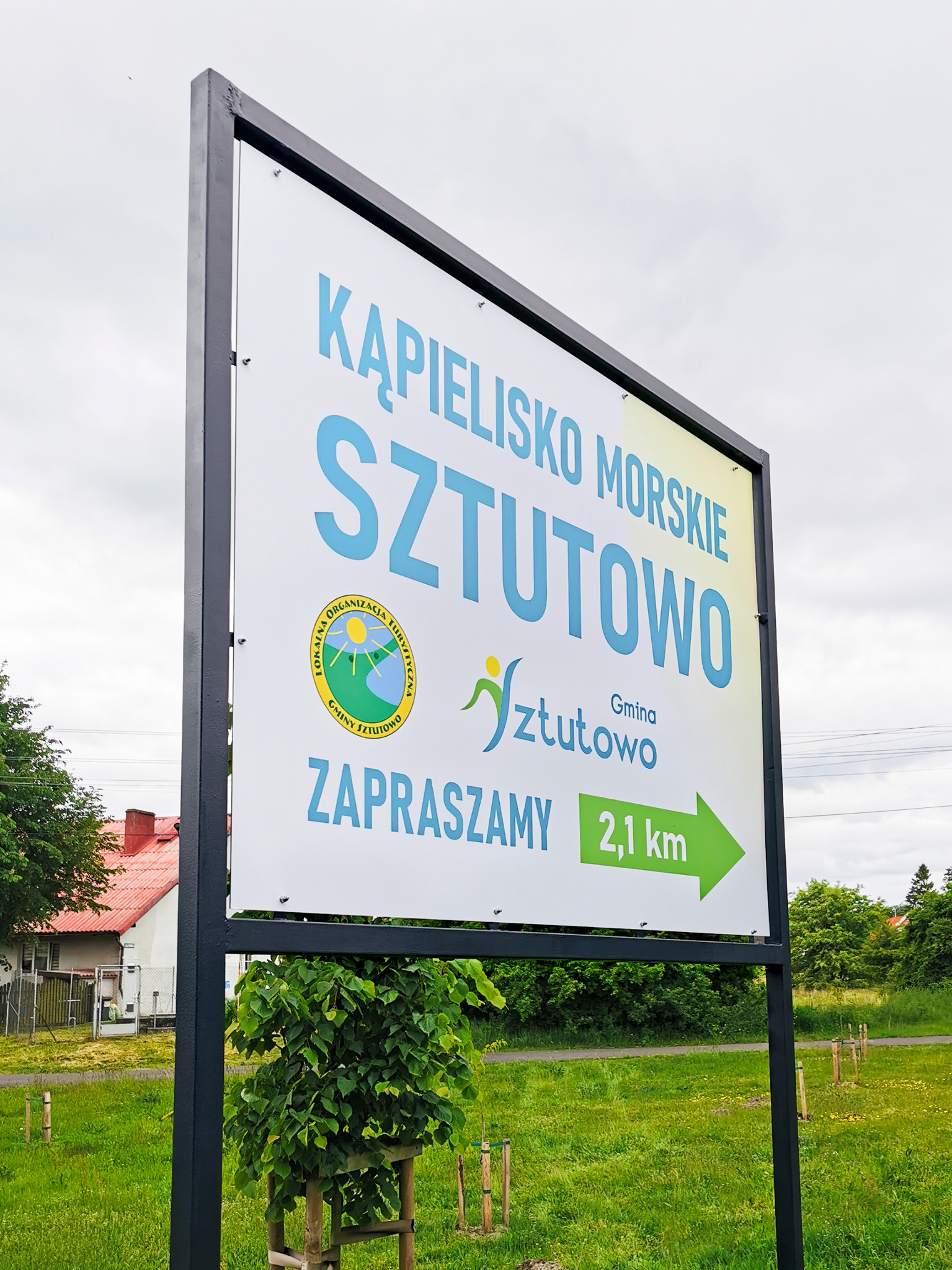 Szyld Lokalnej Organizacji Turystycznej Gminy Sztutowo po odświeżeniu i przemalowaniu stelaża na czarno. Widok z prawej strony po montażu, zieleń w tle.