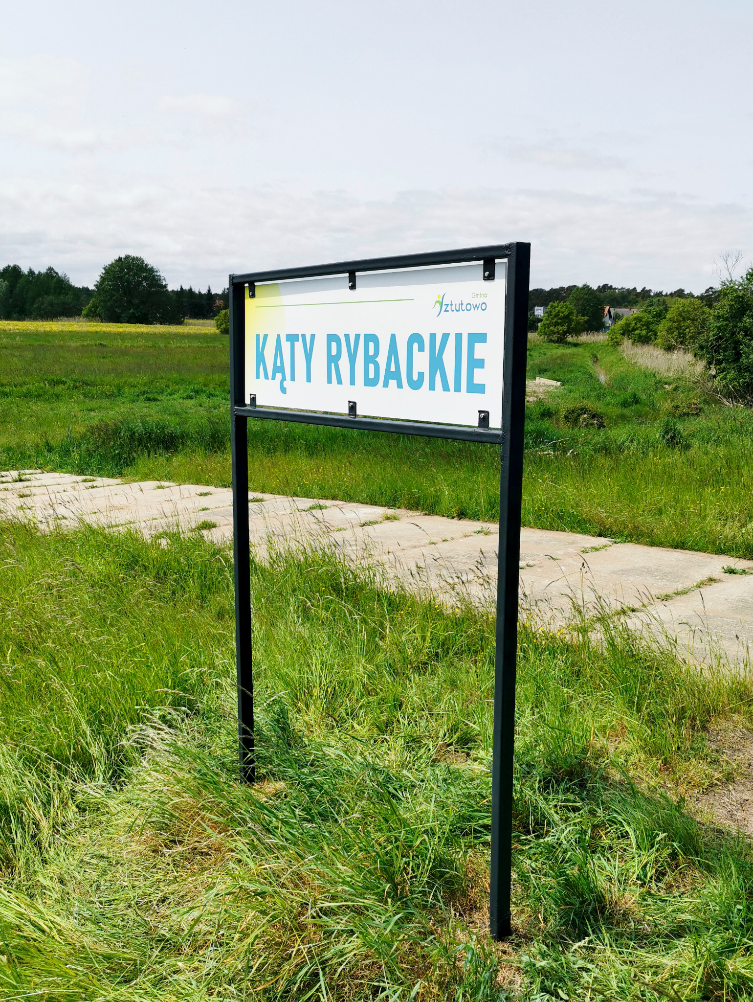 Szyld Lokalnej Organizacji Turystycznej Gminy Sztutowo z nazwą 'Kąty Rybackie' na czarnym stelażu, umieszczony na tle łąki i ścieżki. Odświeżony wygląd.