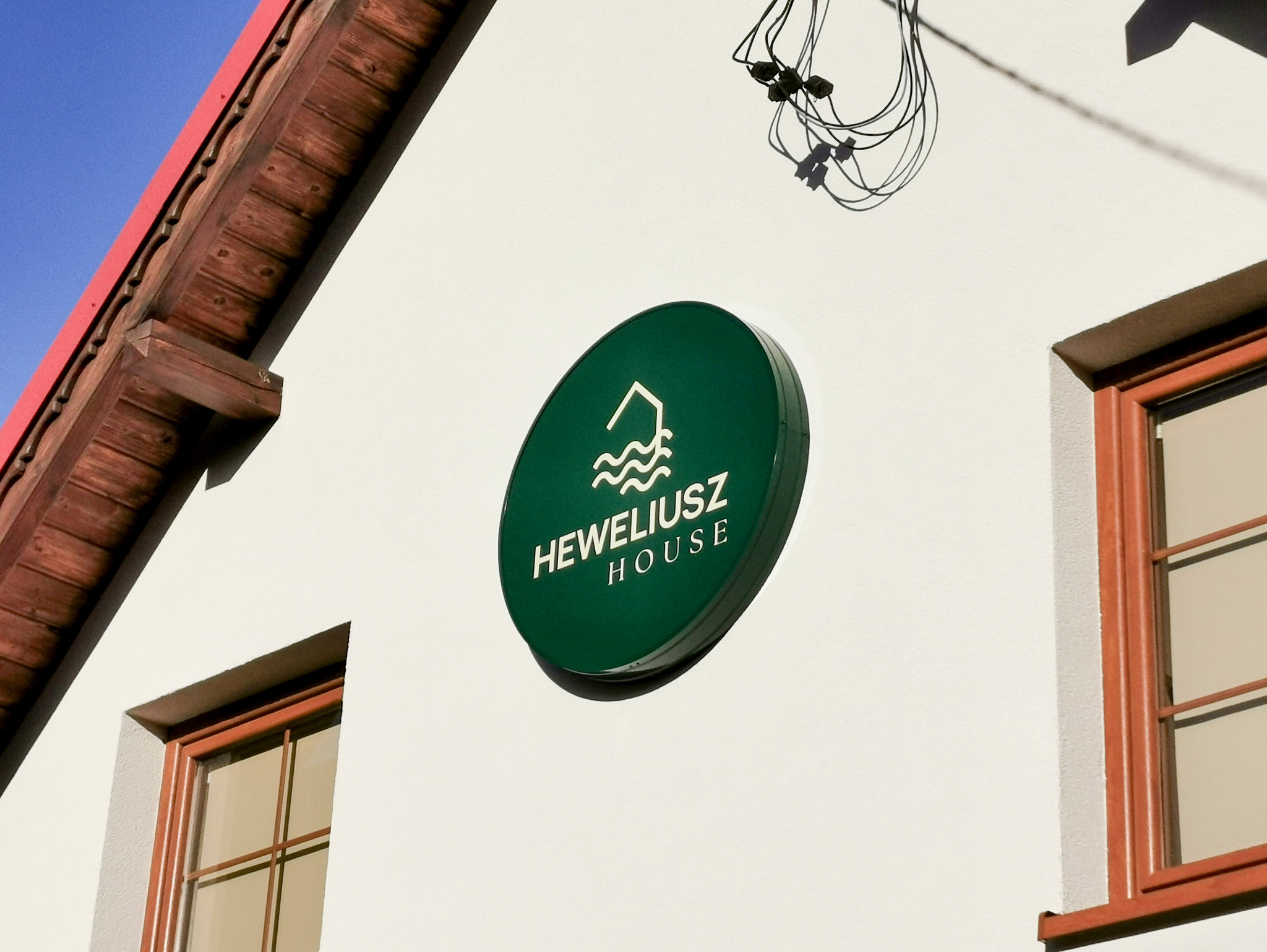 Okrągły, podświetlany, ciemnozielony kaseton z białym logo i napisem 'Heweliusz House' zamontowany na białej ścianie budynku. Widoczne okna z brązowymi ramami.