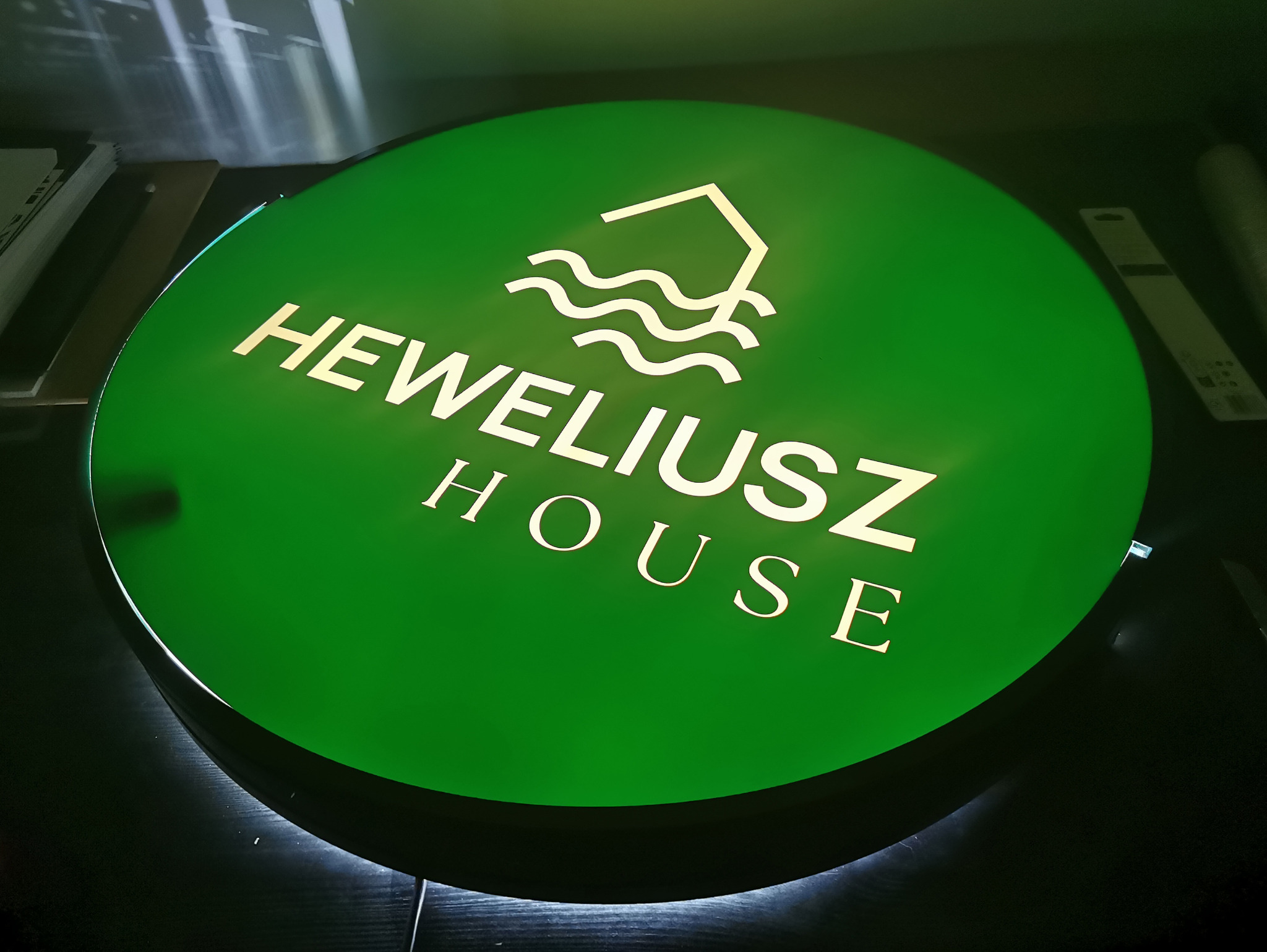 Okrągły, podświetlany kaseton reklamowy w kolorze zielonym z logo i napisem 'Heweliusz House', sfotografowany w studiu podczas testowania oświetlenia.