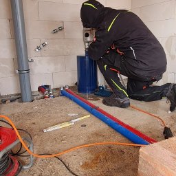 Eko-KlimaWent Bobowicz - Pracownik w kombinezonie wierci otwór w ścianie z betonu komórkowego za pomocą wiertnicy diamentowej. Widoczne rury, przewody i narzędzia na podłodze.