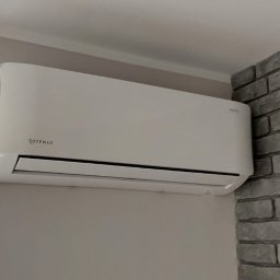 Eko-KlimaWent Bobowicz - Biały klimatyzator Rotenso zamontowany na szarej ścianie, obok okna i ściany z szarej cegły dekoracyjnej. Widok z boku, oświetlenie naturalne.