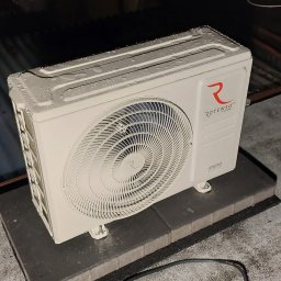 Eko-KlimaWent Bobowicz - Biała jednostka zewnętrzna klimatyzacji Rotenso, pokryta kroplami, umieszczona na szarym podłożu na zewnątrz budynku. Widoczne elementy dachu i przewód zasilający.