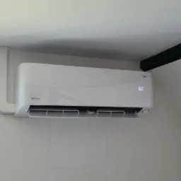 Biały klimatyzator Rotenso Mirai z widocznym wyświetlaczem temperatury 16 stopni Celsjusza, zamontowany na białej ścianie w pomieszczeniu z oknem i ciemną belką stropową.