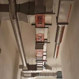 Widok z dołu na system kanałów wentylacyjnych pod sufitem, niektóre z zabezpieczeniami z folii, w tle okno z zabezpieczeniem.